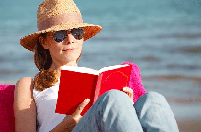 woman wearing reading sunglasses at the beach while reading a book
