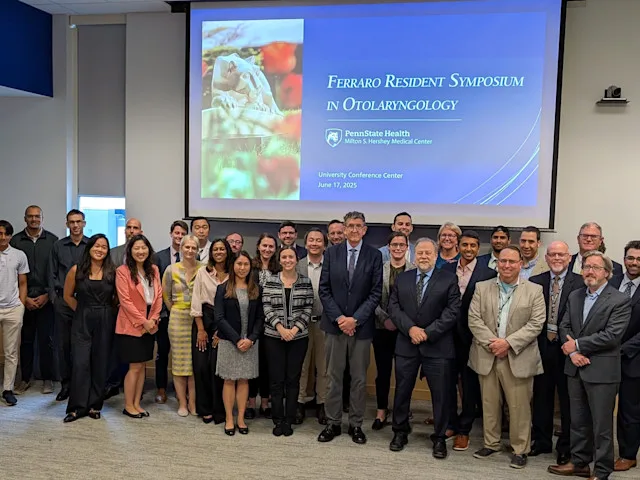 A large group poses in front of projected slide reading “Ferraro Resident Symposium in Otolaryngology” at research conference