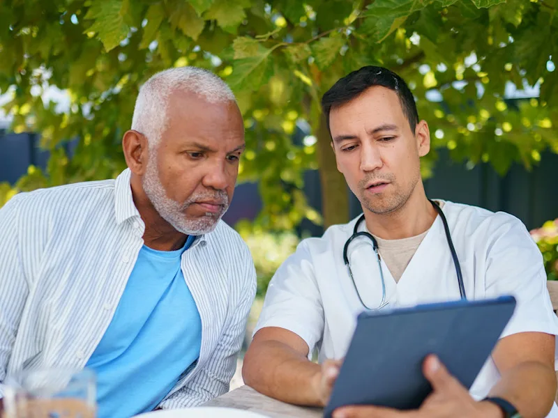 A man with a stethoscope around his neck shows another man something on a tablet in an outdoor setting