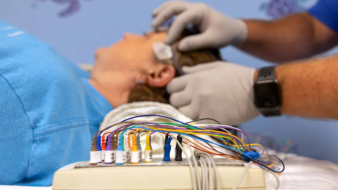A patient lays on a hospital bed, a technician places sensors on her head. A control box with wires sits in the foreground.
