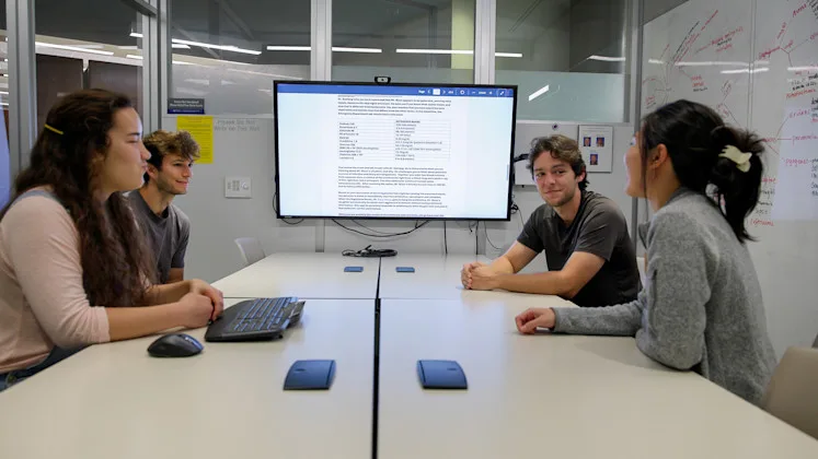 Four students, two at either side of a table, in a study room with a screen at the front