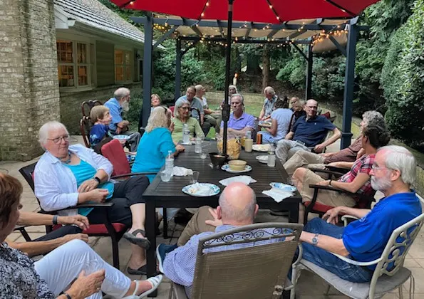 A group of emeritus faculty members sit around an outdoor backyard table under pergolas
