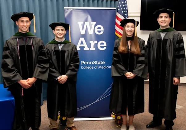 Four students in graduation regalia flanking a We Are Penn State College of Medicine banner