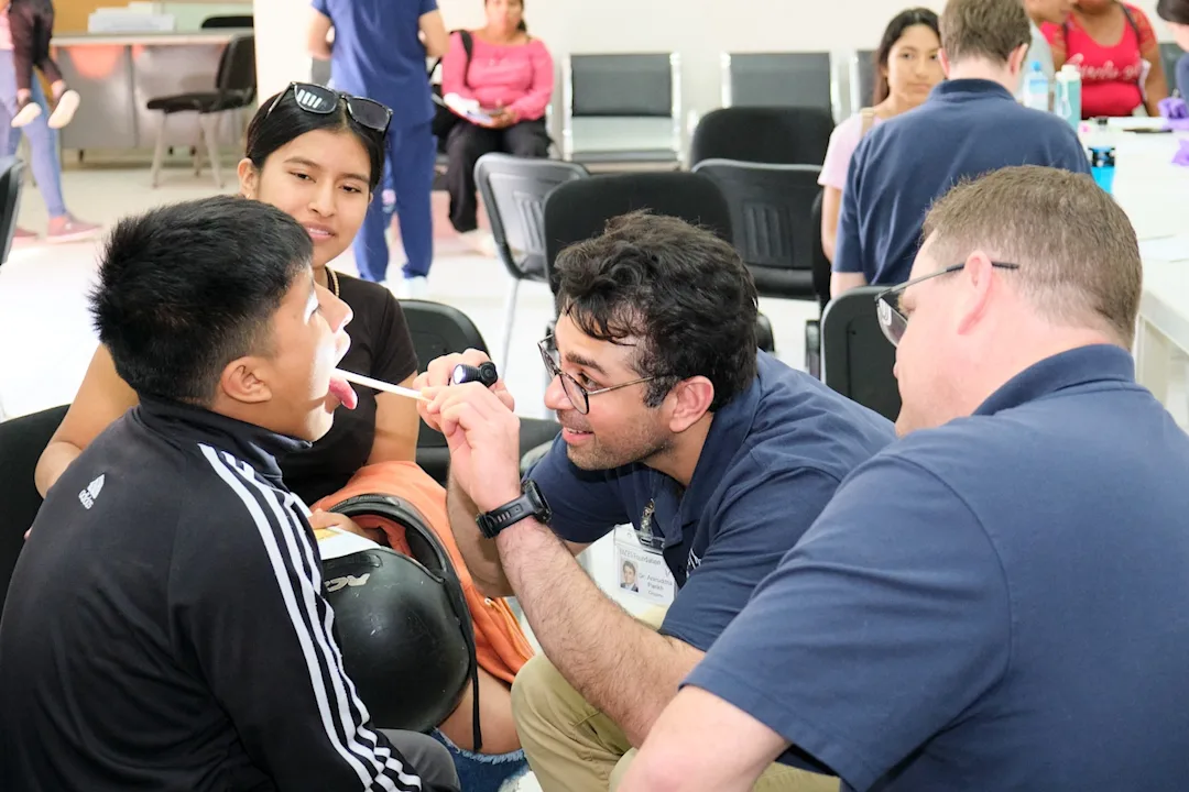 A healthcare professional examines a boy's mouth with a tongue depressor while others wait in the background.