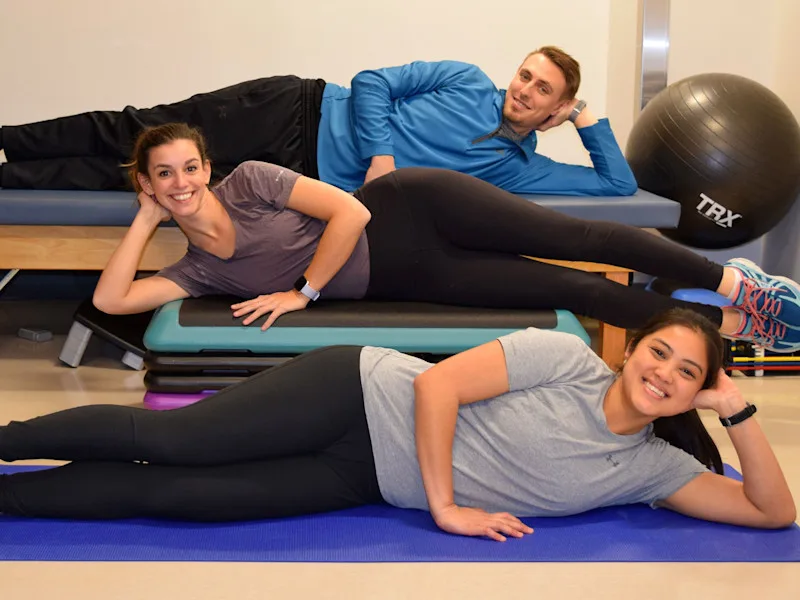 Three people in fitness attire are seen laying on their sides in a workout space.
