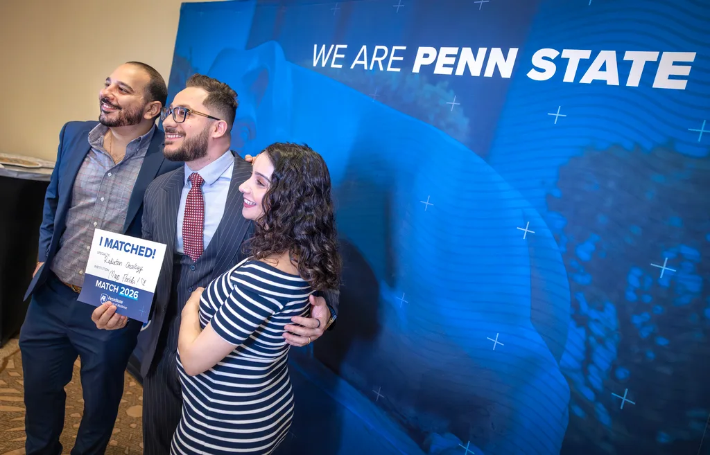 Fadi Samaan, flanked by loved ones, holds a sign with his match information and stands in front of a We Are Penn State sign
