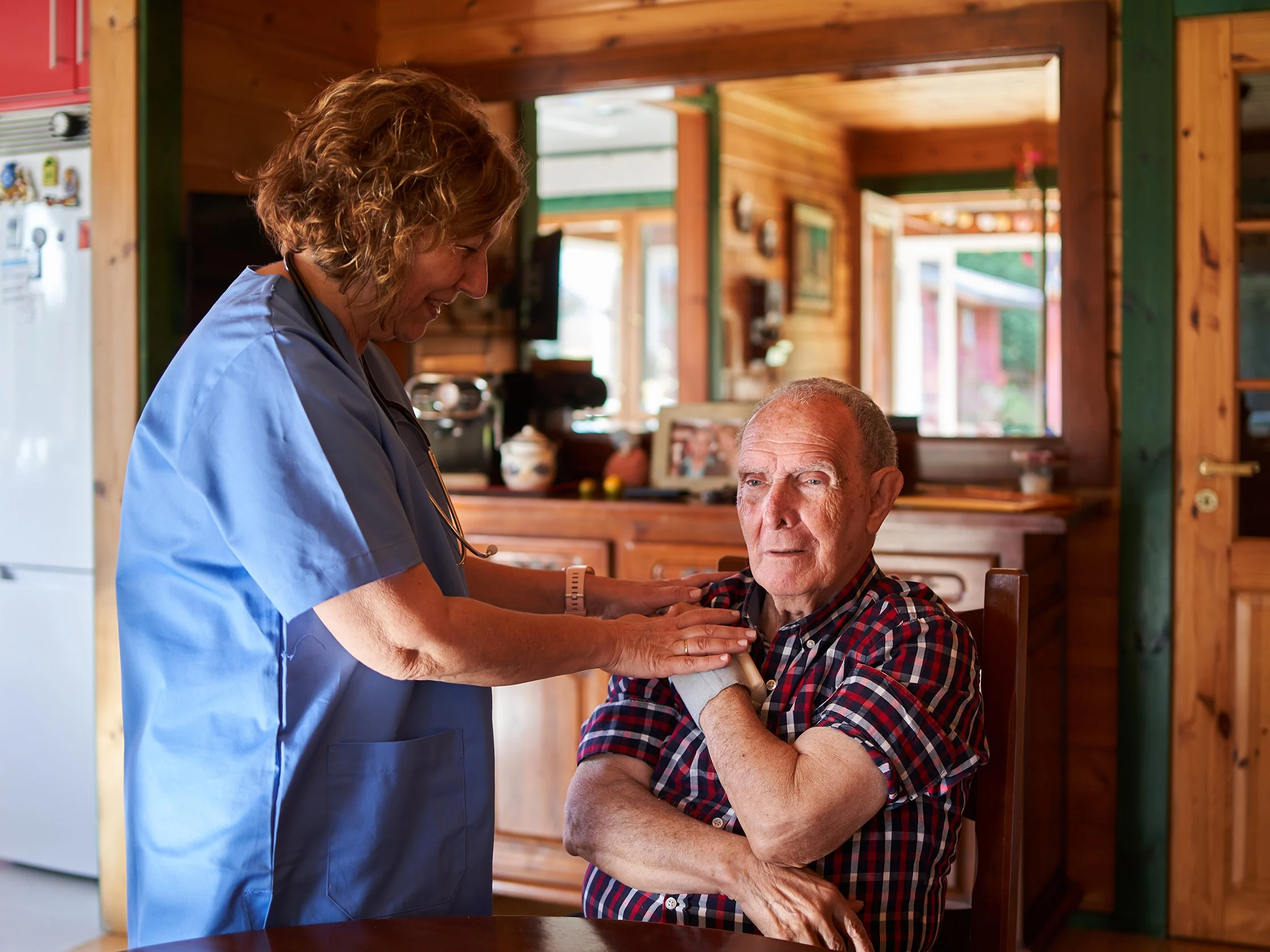 A woman in medical scrubs checks the shoulder of an older man who is sitting in his kitchen