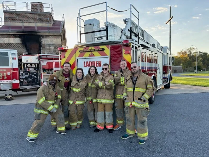 Seven Emergency Medicine residents stand behind a fire truck wearing turnout gear