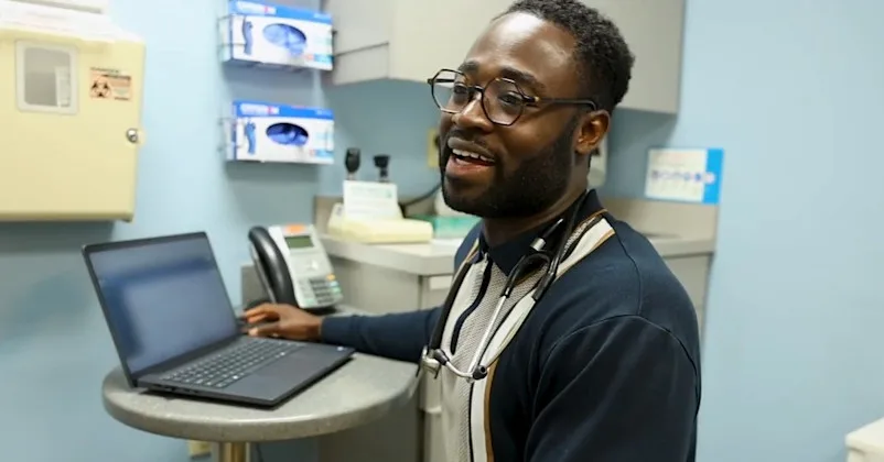 A resident smiles and talks with someone off-screen with a laptop on a small table next to him