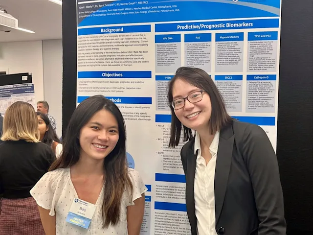 Two medical students stand in front of a research poster at a medical or scientific conference, beside their displayed poster