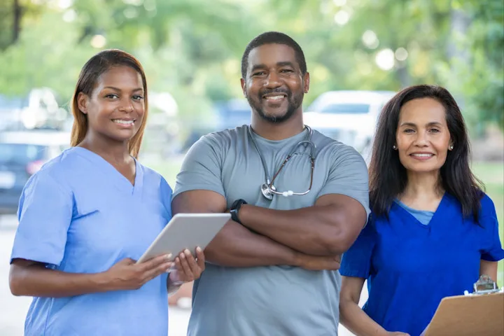 Three health providers wearing scrubs; two women on the flanks hold clipboards or tablets; man in center has a stethoscope