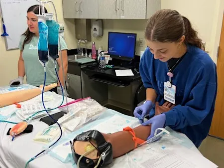 Student practicing IV insertion on a simulation arm while another student observes in a clinical skills room.