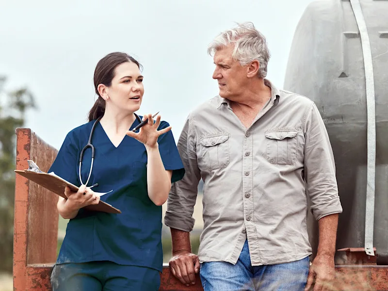 A woman wearing medical scrubs and a stethoscope and holding a clipboard gestures while talking to a man