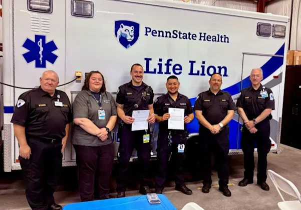 Six people in front of a Penn State Health Life Lion vehicle; two fellows in the center are holding awards