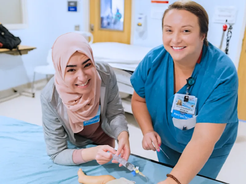 Two smiling medical trainees practice IV insertion on a newborn manikin in a clinical simulation room