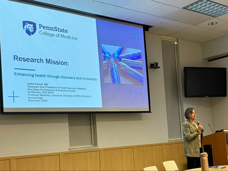 Dr. Leslie Parent holds a microphone and stands next to a lectern, with a large screen projecting a slide to her left