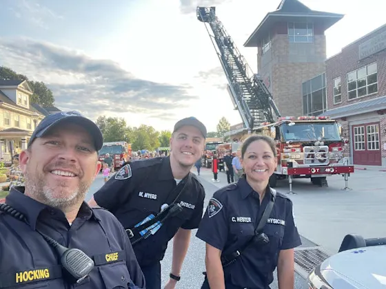 Three fellows take a selfie during National Night Out with a fire truck, ladder up, in the background