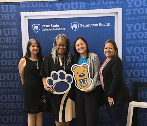 Four women in front of Penn State Health and College of Medicine-branded frame and holding paw print and Nittany Lion props