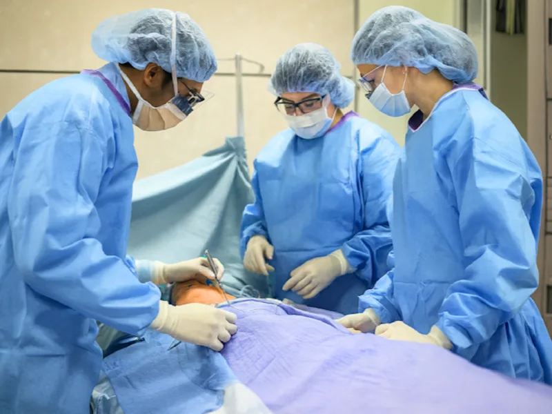 Three healthcare professionals in full surgical attire performing a simulated operation on a medical training manikin.