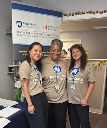 Three women in Penn State College of Medicine shirts next to their informational table with a CAHE-RUC pulldown banner