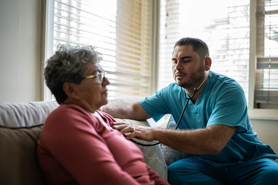 A man uses a stethoscope on a woman on a couch inside a home