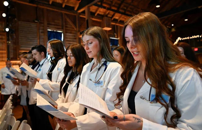 A row of incoming physician assistant students wearing white coats read from a program