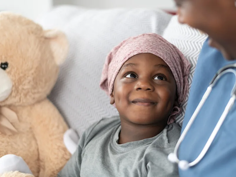 A young patient with head covering sits with a teddy bear off to the side and looks up at a clinical provider