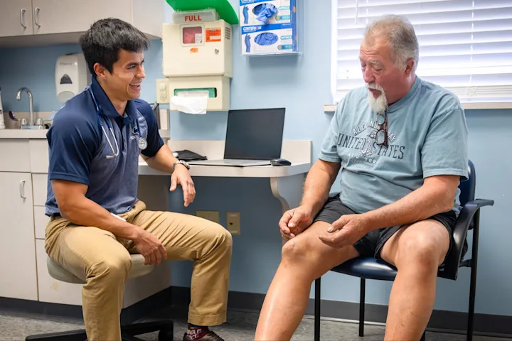 3+ MD student Kyle Nerz sits in a clinical exam room and talks with a patient who is gesturing toward his knee