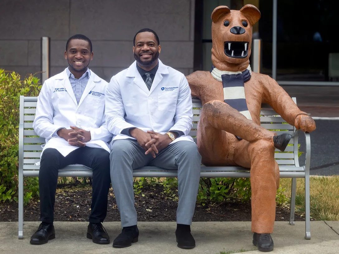 Two fellows wearing physicians' white coats sit on a bench with a Nittany Lion mascot statue