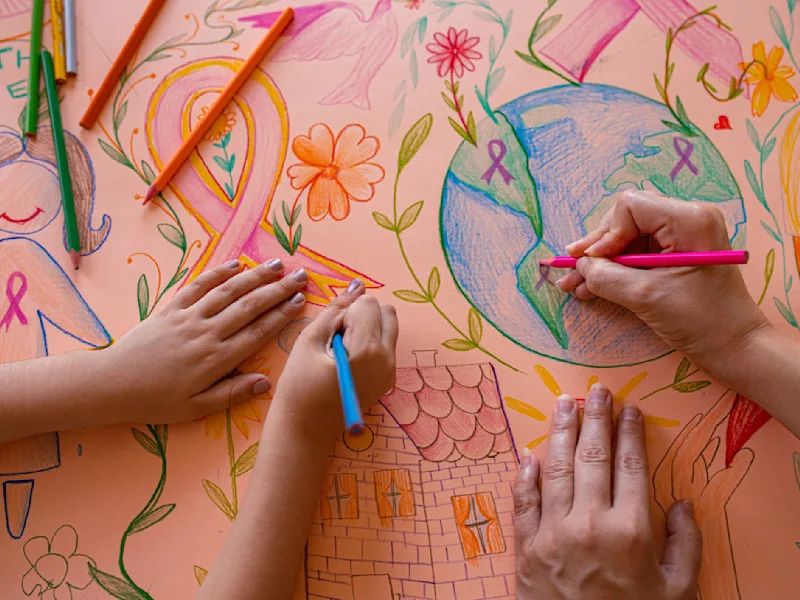 Set of adult hands and set of child hands using colored pencils to color a picture that includes a ribbon, earth and building
