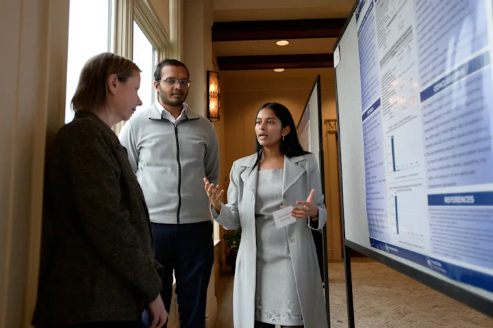 Three people stand beside a research poster at a medical conference, discussing poster content on a presentation board