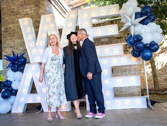 A graduating medical student flanked by her mom and dad in front of a We Are sign