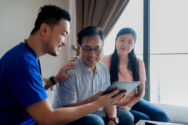 A man in blue scrubs and stethoscope with his hand on another man's shoulder while he shows him a tablet; a woman looks on