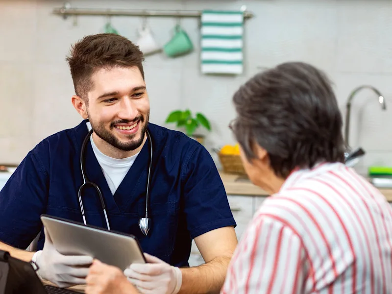 A man with a stethoscope around his neck smiles and holds a tablet while talking to an older woman