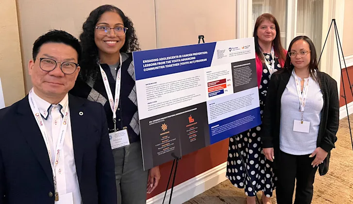Four conference attendees with lanyards stand beside a research poster titled 'Engaging Adolescents in Cancer Prevention'