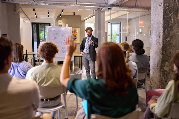 A man in a suit presents to a classroom with a flip chart, while a participant raises their hand.