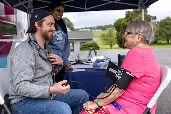 A medical student takes the blood pressure of a community member outdoors while another student looks on
