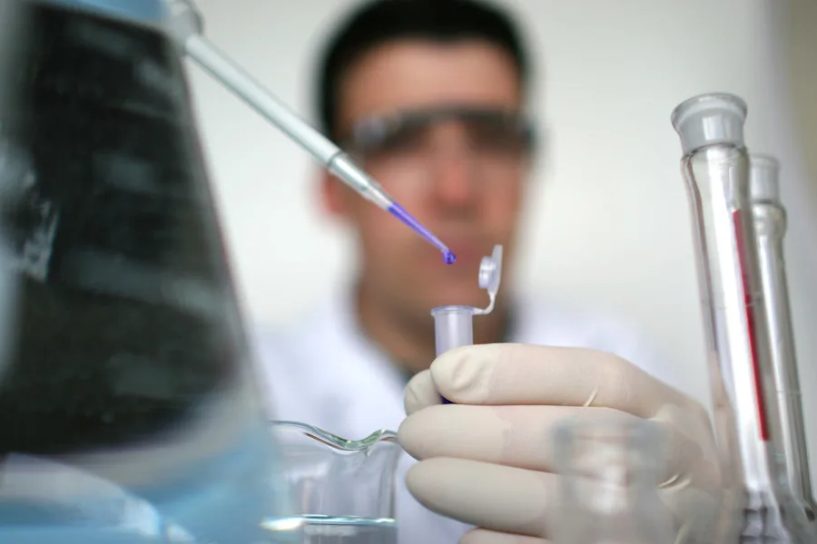 A researcher holds a test tube while dropping liquid from a dropper; the person's face is blurred in the background
