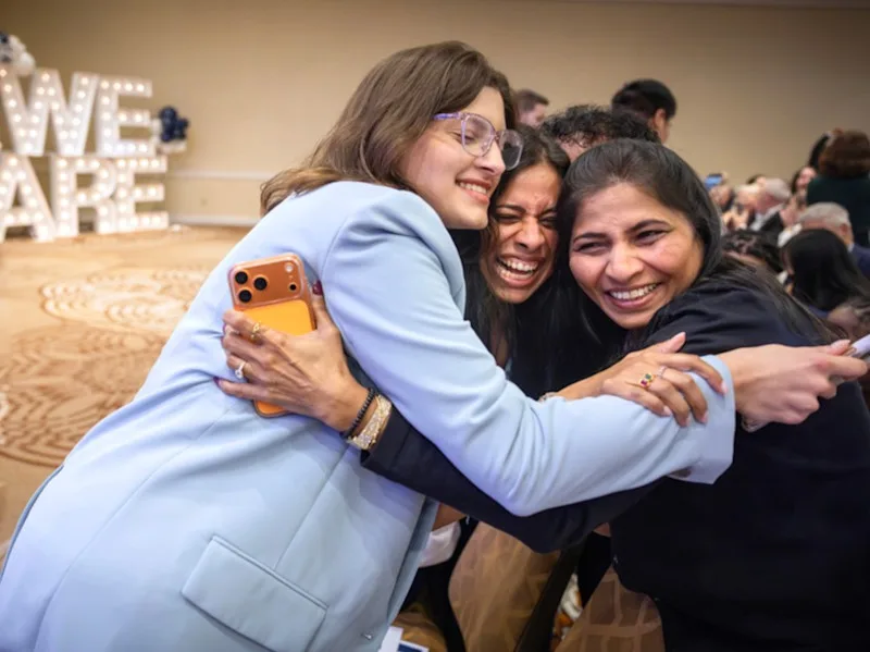 With a light-up We Are sign in the background, three people hug and smile after MD students found out their matches
