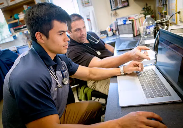A student with a stethoscope around his neck looks at a laptop while a faculty mentor points in a clinical setting