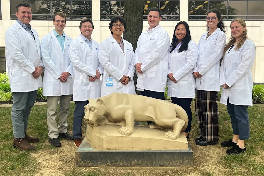 5 residents in physician white coats pose behind the Nittany Lion statue in a courtyard