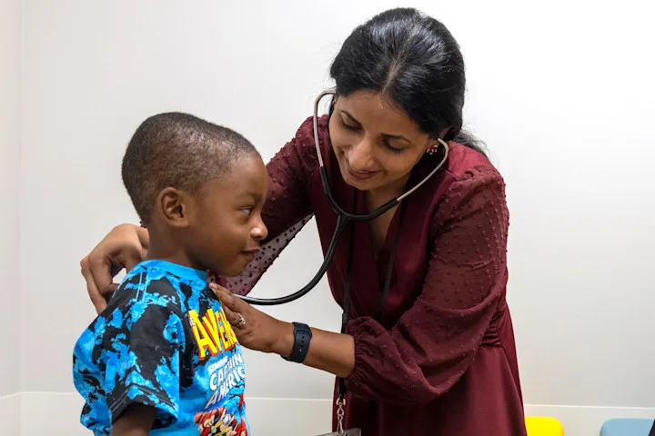 A physician uses a stethoscope to check the vitals of a young boy who is sitting on an exam table
