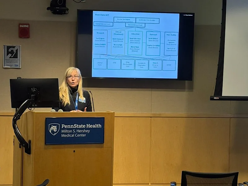 A presenter speaks from behind a lectern while a slide is projected behind and to her left
