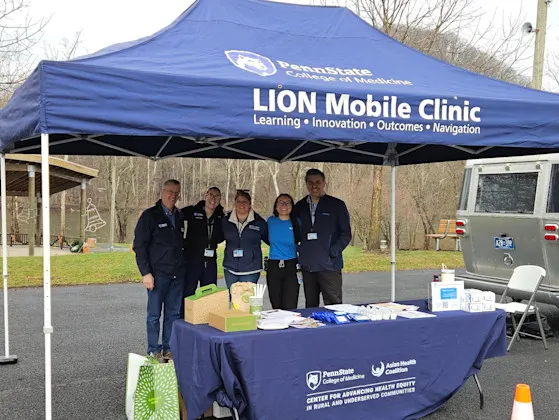 Staff under a LION Mobile Clinic tent behind a table with a CAHE-RUC tablecloth at an outdoor community health event.