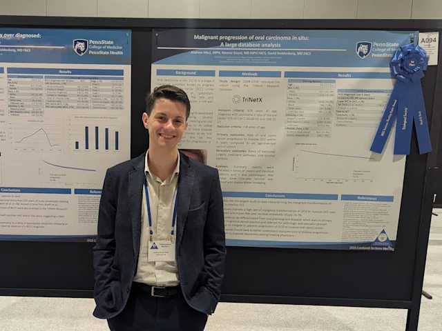 An individual stands in front of a research poster at a conference, with a blue first‑place ribbon attached to the poster