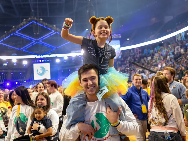 A young girl who benefits from Four Diamonds pumps her fist while sitting on a college student's shoulders at THON.
