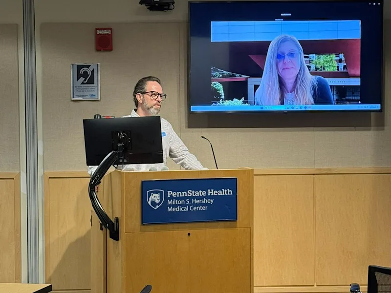 Yuval Silberman stands behind a lectern while introducing Camelia Kantor, who is on a screen to his left.