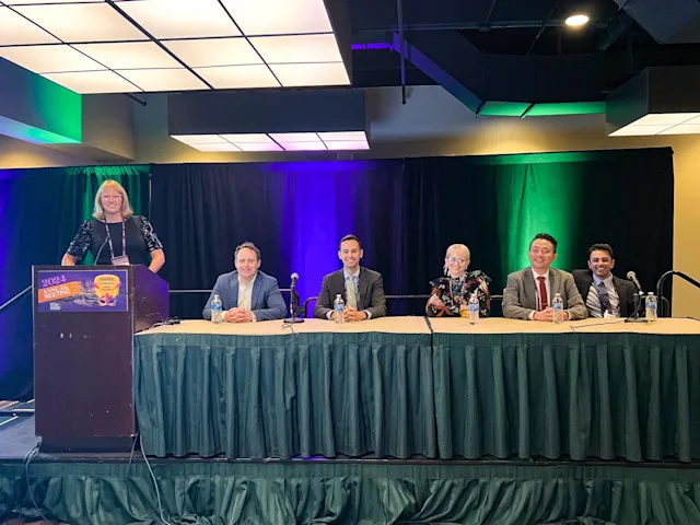 A team sits behind a long table on a conference stage at a medical conference, with one person standing at a podium