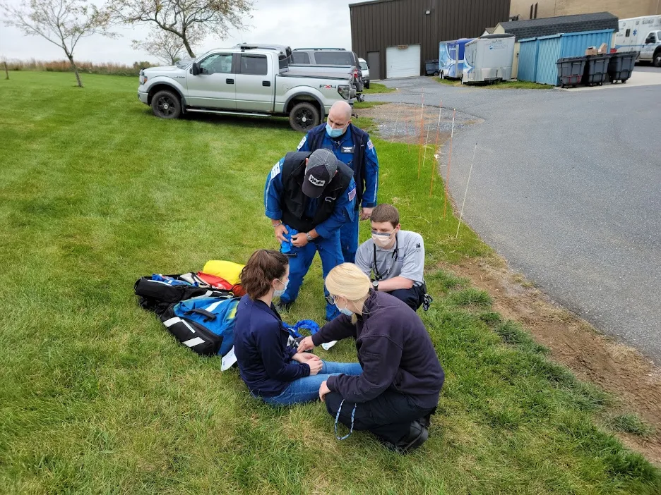 In an EMS Fellowship training exercise, two people kneel and two stand near a patient who is sitting on the ground