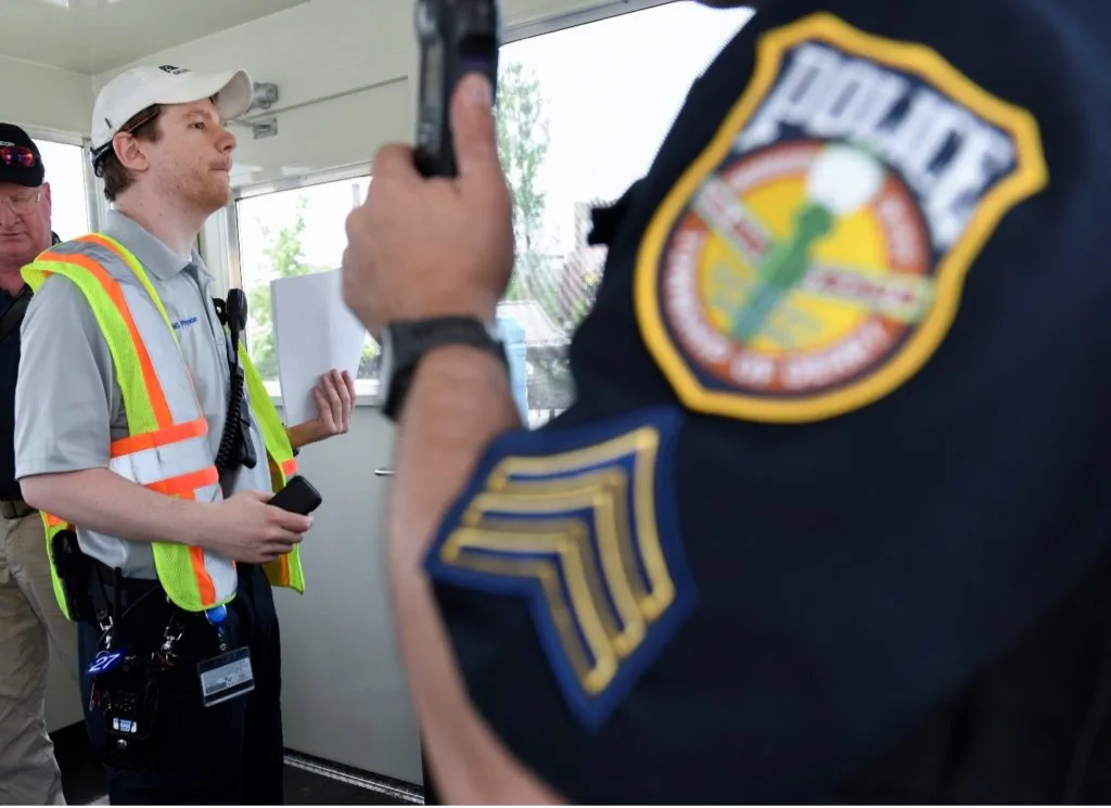 A police arm badge in the foreground while a man in an orange vest holds papers.